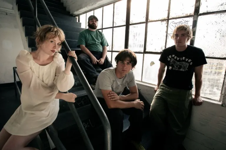 Indie rock band Swapmeet four members pose in an industrial stairwell next to large grid windows, featuring a woman in a white dress on the handrail.
