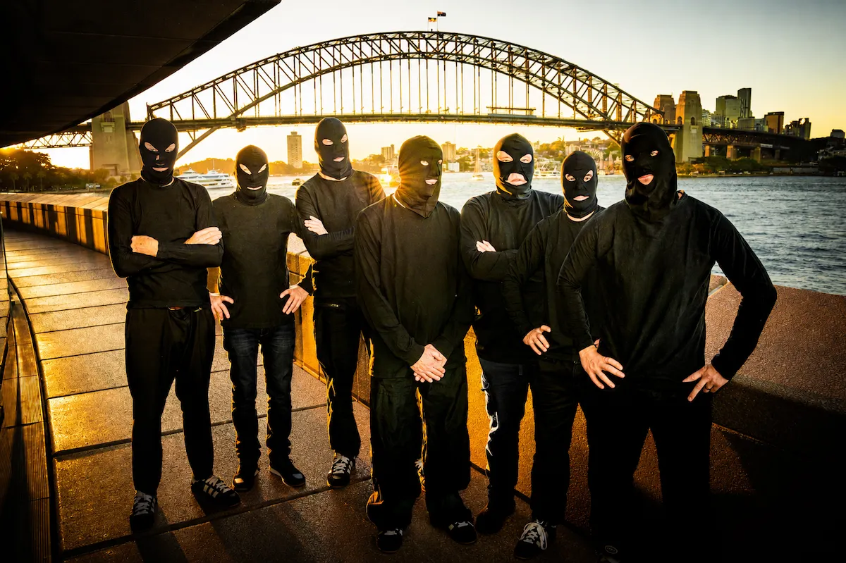 Promotional photo of Australian rock band TISM wearing balaclavas by Sydney Harbour at sunset, with the Harbour Bridge behind them.