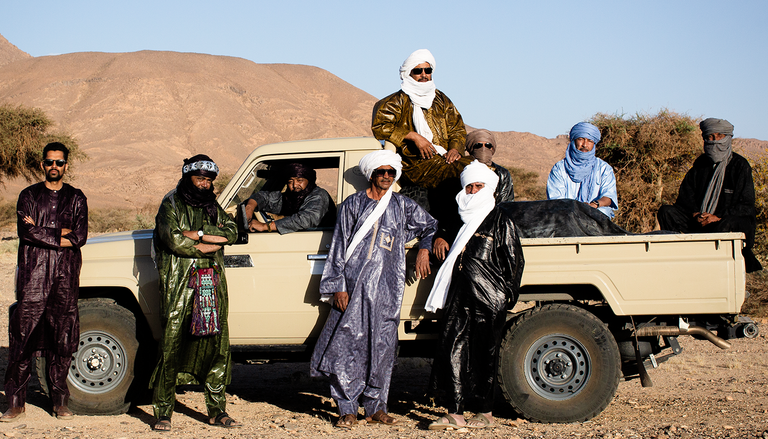 Tuareg band Tinariwen posing around a pickup truck in traditional garments and headwraps against a desert landscape.