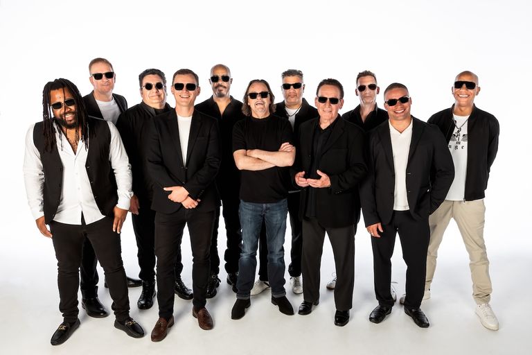 Reggae band UB40 group photo featuring band members wearing dark sunglasses and black and white outfits standing against a plain white background.
