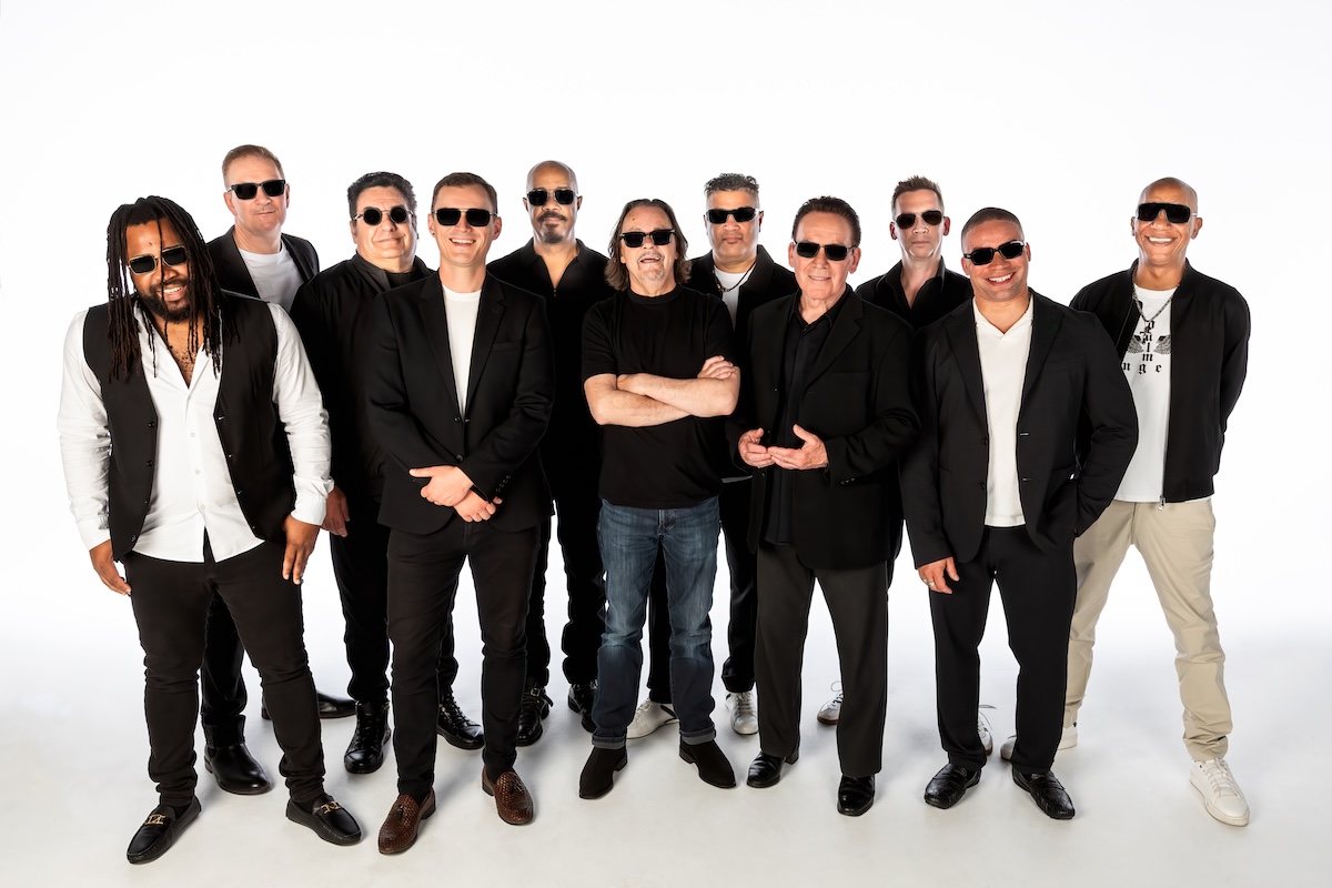 Reggae band UB40 group photo featuring band members wearing dark sunglasses and black and white outfits standing against a plain white background.