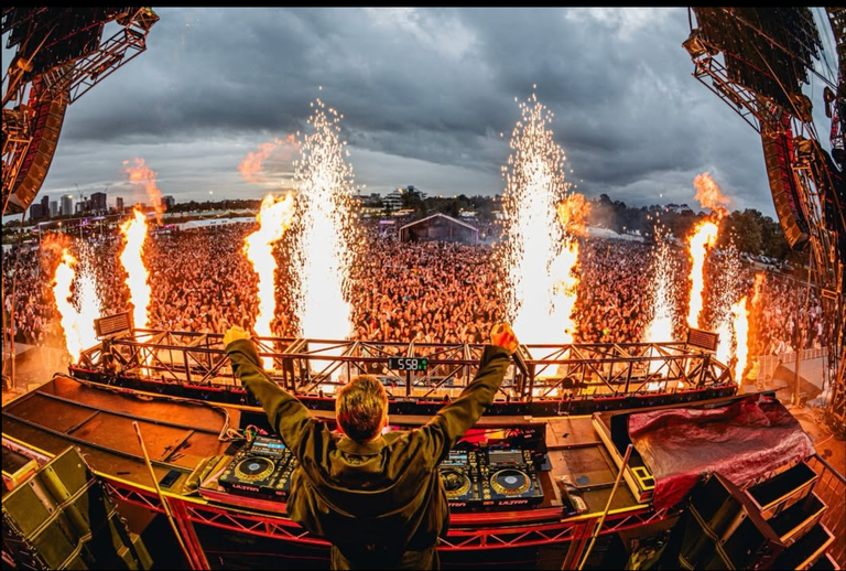 A DJ performs on stage with arms raised in front of a massive crowd with pyrotechnics and flames at Ultra Music Festival.