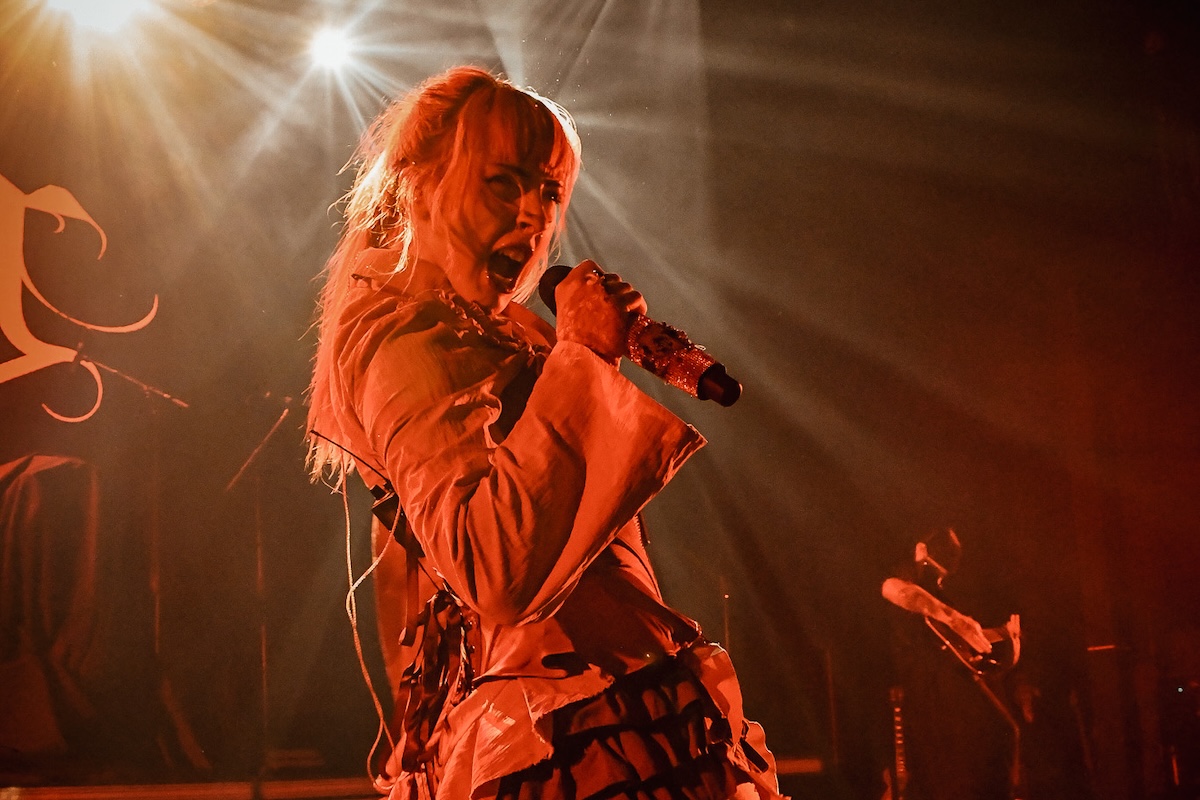 VANA singing passionately into a microphone on stage under warm, dramatic orange lighting during an energetic live musical performance.