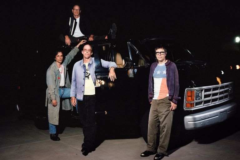 Promotional photo of American rock band Weezer posing around a black van at night.
