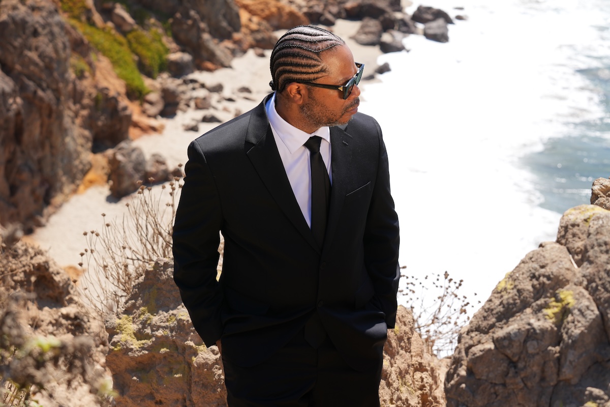 Rapper Xzibit wears a black suit, sunglasses, and braided hair while standing on rocky coastal cliffs looking toward the ocean waves.