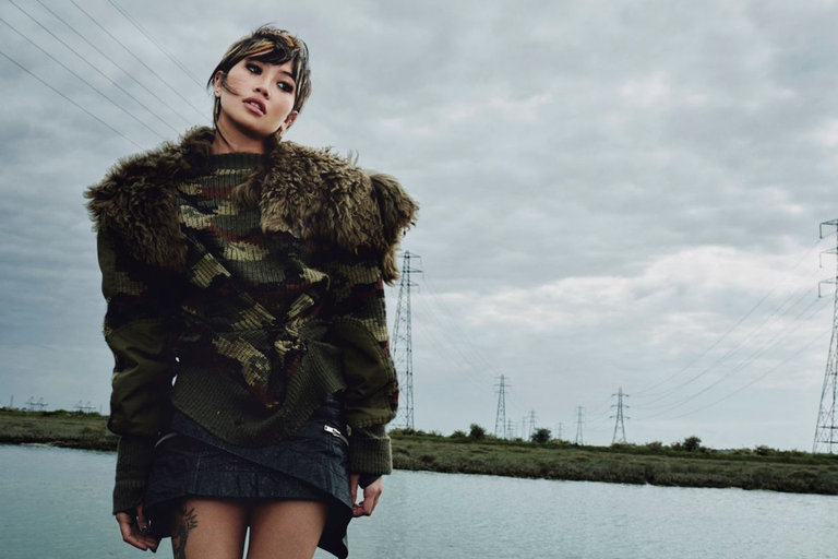 Promotional portrait of indie musician beabadoobee wearing a fur-trimmed knit jacket and standing outdoors near power lines under a cloudy sky.