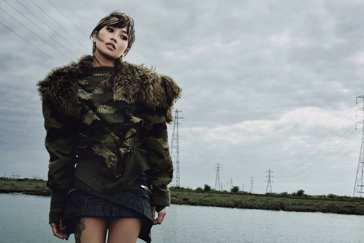 Promotional portrait of indie musician beabadoobee wearing a fur-trimmed knit jacket and standing outdoors near power lines under a cloudy sky.