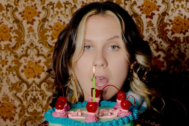 Singer Grace Robinson looks over a retro-decorated birthday cake topped with cherries and a single lit candle, set against wallpaper.
