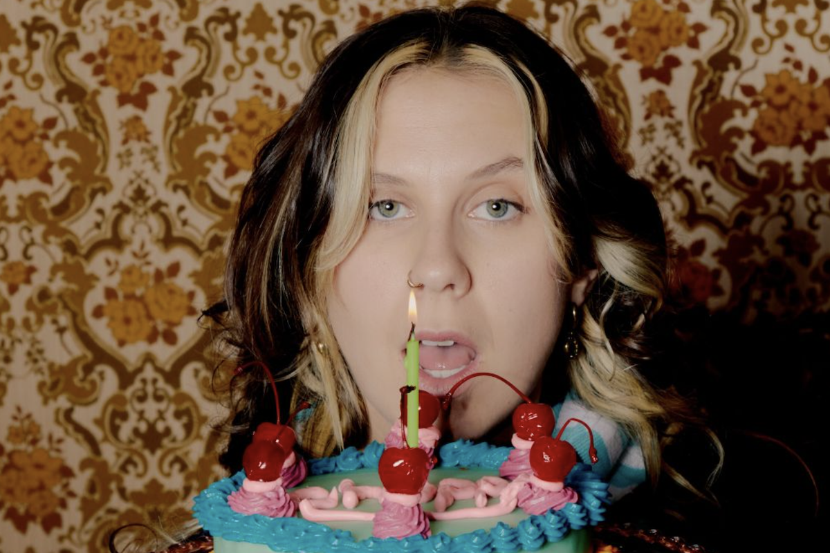 Singer Grace Robinson looks over a retro-decorated birthday cake topped with cherries and a single lit candle, set against wallpaper.