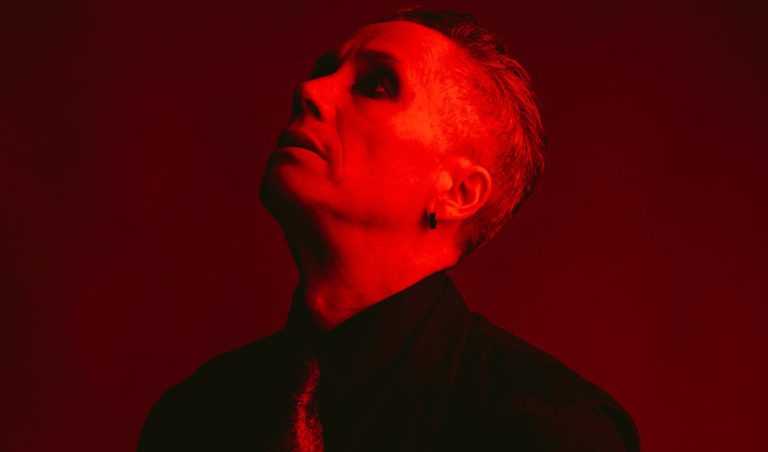 Dramatic portrait of musician and performer iOTA bathed in intense red light, looking upward in a black shirt and tie.