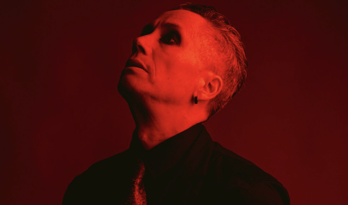 Dramatic portrait of musician and performer iOTA bathed in intense red light, looking upward in a black shirt and tie.