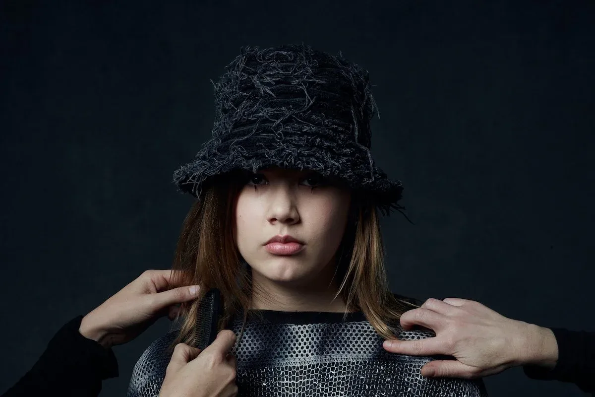 Maple's Pet Dinosaur portrait featuring a person in a textured dark bucket hat as hands adjust hair and clothing against a dark background.