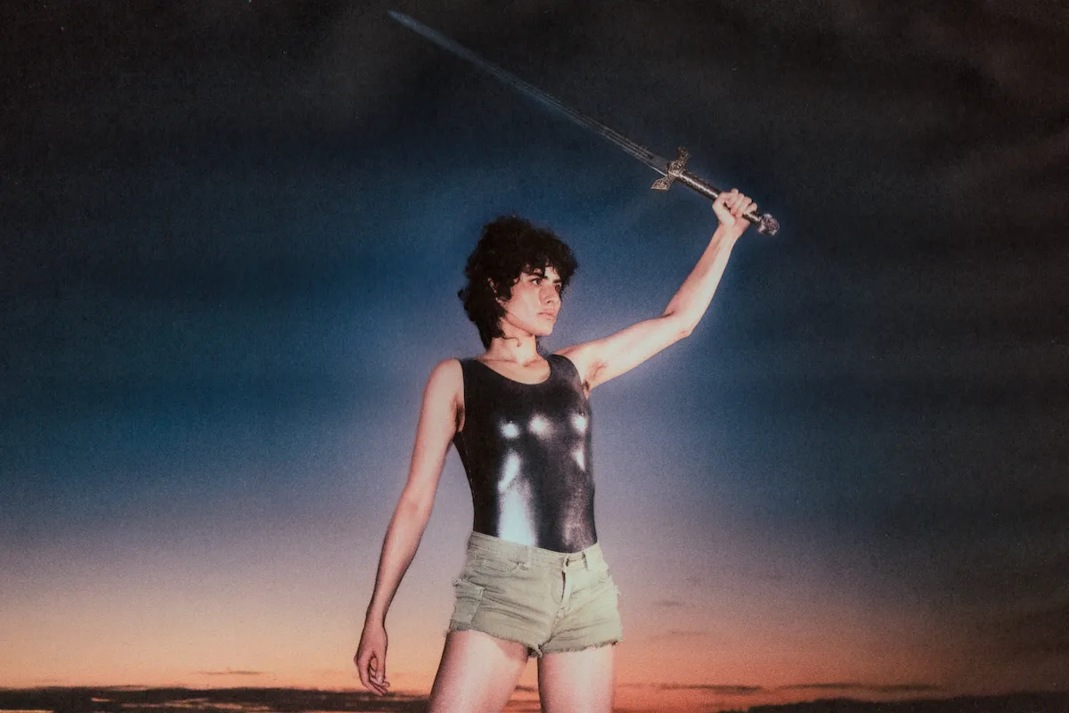 Australian art-pop artist Montaigne wearing a metallic tank top and holding a sword raised against a twilight sky.