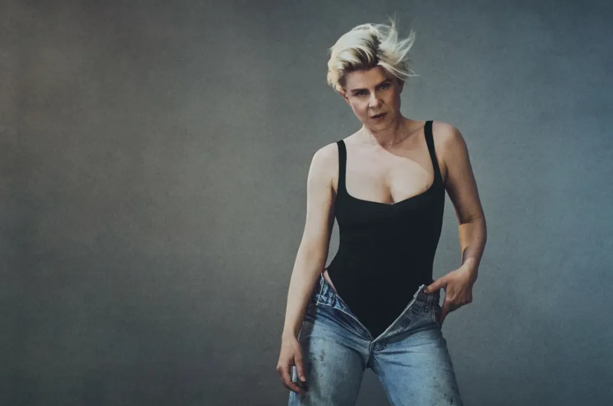 Pop singer Robyn posing in a black bodysuit and unbuttoned jeans with blonde hair against a grey backdrop.