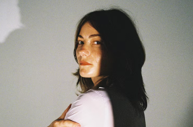 Promotional portrait of indie folk singer-songwriter Rowena Wise looking over her shoulder in warm light against a neutral backdrop for music press.