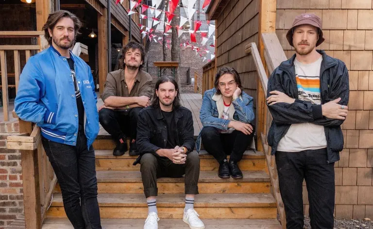 Indie soul-pop band The Wanderers posing casually outdoors on wooden steps in front of a shingled building with red and white bunting.