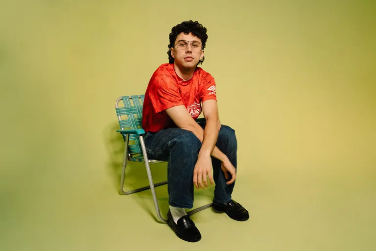 Australian DJ and producer Young Franco sitting on a green lawn chair in a red sports jersey and jeans against a solid lime green backdrop.